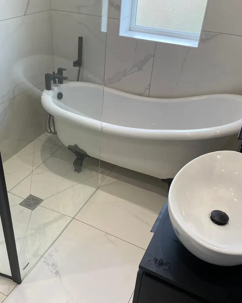 Walk-in shower with a black-framed glass screen and floor drain beside a roll-top bath